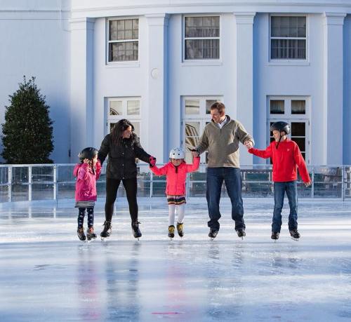 A family ice skating together, holding hands on a rink, with a white building in the background.