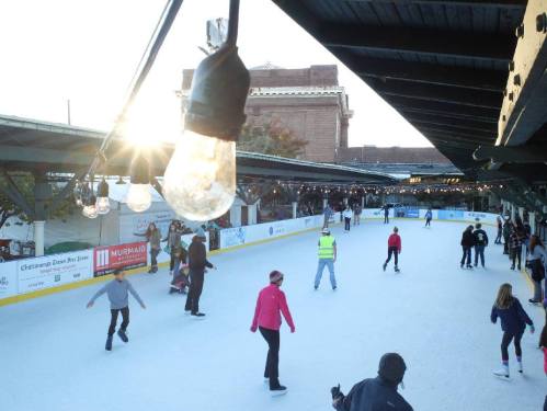 A bustling ice skating rink with people skating under string lights, set against a sunset backdrop.
