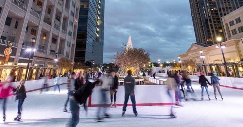 A bustling outdoor ice skating rink with people skating, surrounded by festive lights and buildings under a cloudy sky.