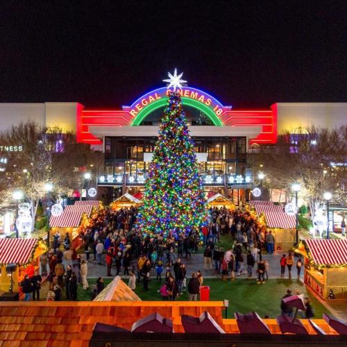 A large, colorful Christmas tree stands in front of a cinema, surrounded by festive stalls and a crowd of people.