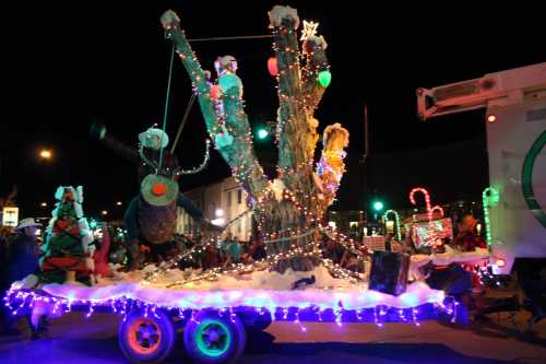 A festive parade float decorated with lights, featuring a whimsical tree and holiday characters against a night backdrop.