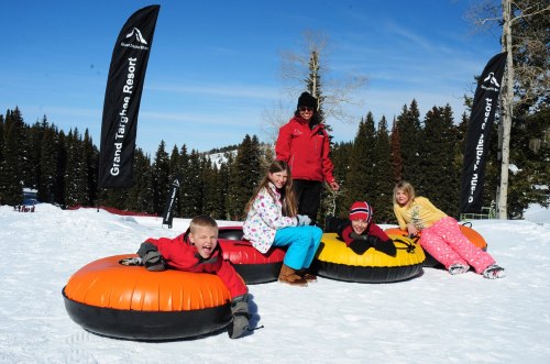 A group of children and an adult enjoy snow tubing at a winter resort, surrounded by snowy trees and flags.