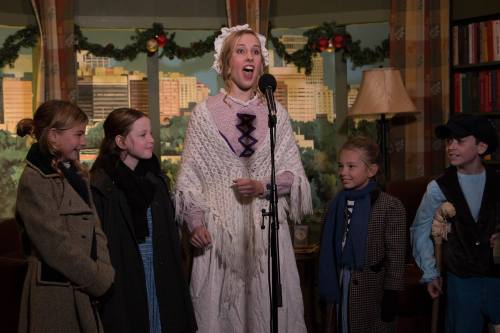 A performer in period costume sings into a microphone, surrounded by four children in vintage attire.