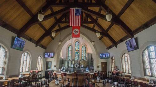 Interior of a restaurant with high wooden ceilings, large windows, and an American flag hanging above the bar area.