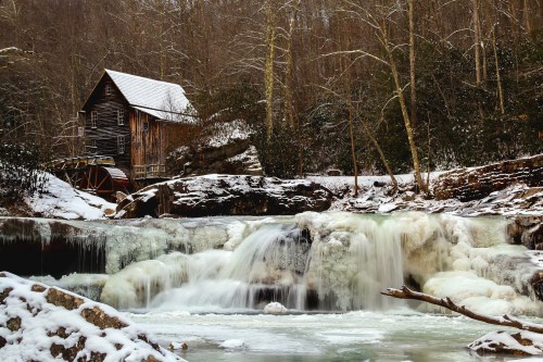 A rustic wooden mill beside a frozen waterfall, surrounded by snow-covered trees in a serene winter landscape.