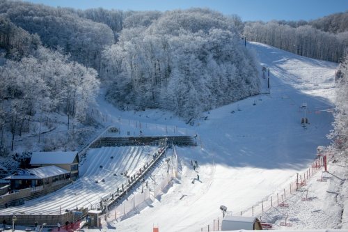 Snow-covered ski slope with trees in the background, a ski lift, and a small building at the base. Clear blue sky.