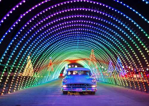 A vintage car drives through a colorful, illuminated archway of lights, surrounded by festive decorations.