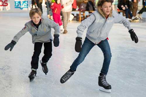 Two boys skate on an ice rink, smiling and enjoying their time, surrounded by other skaters in a festive atmosphere.