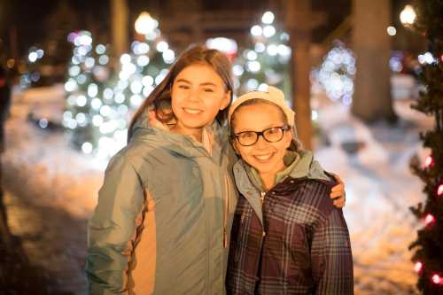 Two smiling girls in winter jackets pose together in a snowy, festive setting with twinkling lights in the background.