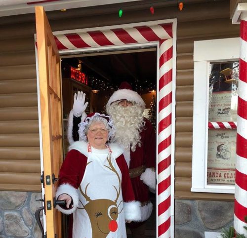 A cheerful Santa and Mrs. Claus stand at a festive door, welcoming visitors with holiday decorations around them.