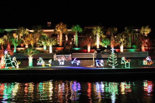 Colorful Christmas lights decorate a waterfront property, featuring festive figures and palm trees against a night sky.
