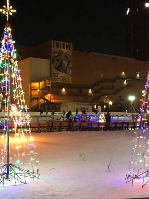 Colorful holiday lights adorn trees in a snowy area, with people skating and a large banner in the background.