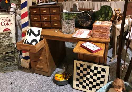 A vintage wooden desk with drawers, surrounded by various antiques, including a clock, a chessboard, and a doll.