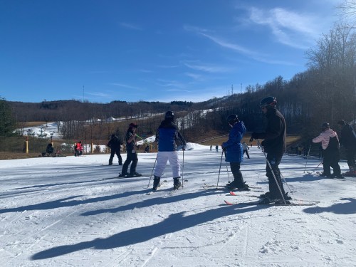 Skiers on a snowy slope under a clear blue sky, with trees and hills in the background.