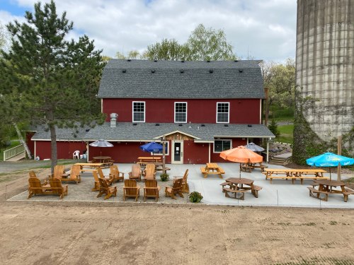 A red barn-style building with outdoor seating, umbrellas, and a gravel area, surrounded by trees and a silo.
