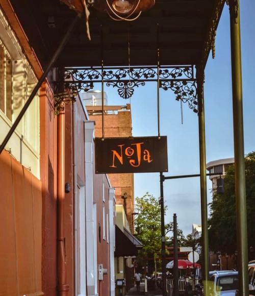 A street view featuring a decorative sign that reads "NJa" hanging from an ornate bracket against a colorful building.