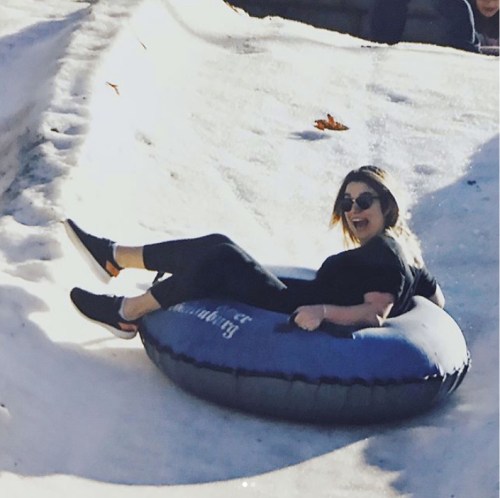 A person joyfully sliding down a snowy slope on a blue inner tube, wearing casual clothes and sunglasses.