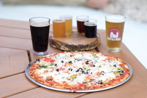A pizza topped with vegetables and cheese, surrounded by various glasses of beer on a wooden table.