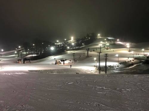A snowy ski slope illuminated by lights at night, with skiers and snowboards visible on the trails.