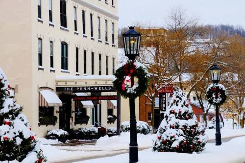 Snow-covered street scene featuring festive wreaths, lampposts, and the Penn-Wells Hotel in a winter setting.