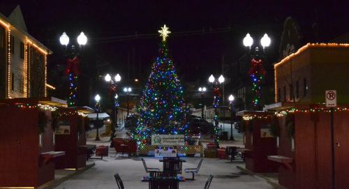 A brightly lit Christmas tree adorned with colorful lights stands in a festive town square at night.
