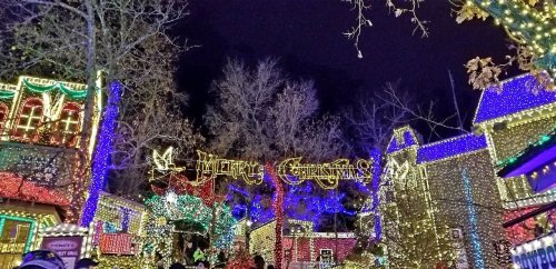 A festive display of colorful Christmas lights adorning trees and buildings at night, with a "Merry Christmas" sign.