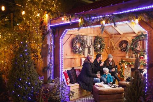 A cozy holiday scene with a family enjoying drinks in a decorated cabin, surrounded by festive lights and greenery.