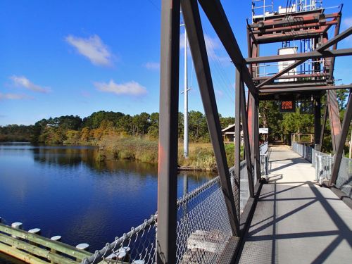 A metal bridge spans a calm river, surrounded by trees and blue skies, with a power line in the background.