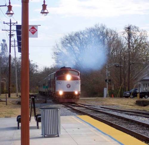 A vintage train approaches a station, with smoke billowing and trees in the background under a clear sky.