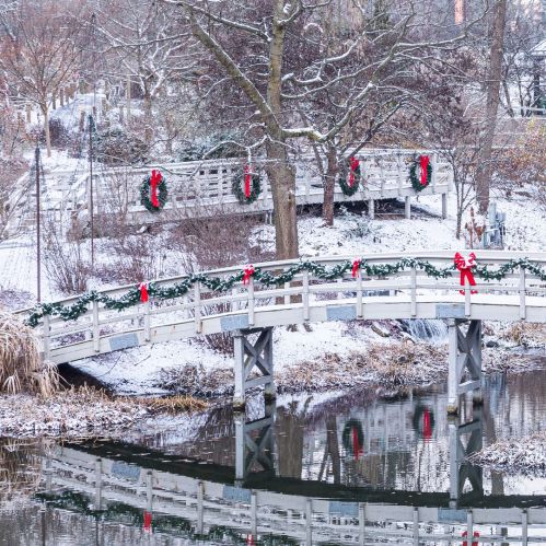 Snow-covered landscape featuring decorated wooden bridges with wreaths and red bows, reflecting in a calm waterway.