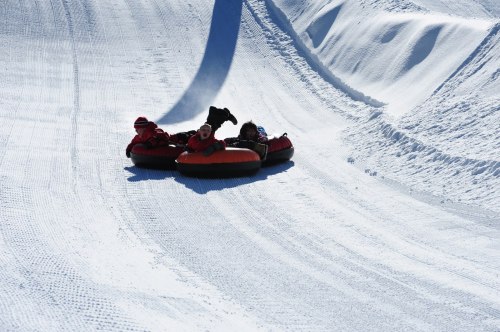 Three people enjoy tubing down a snowy slope, surrounded by pristine white snow and a clear blue sky.