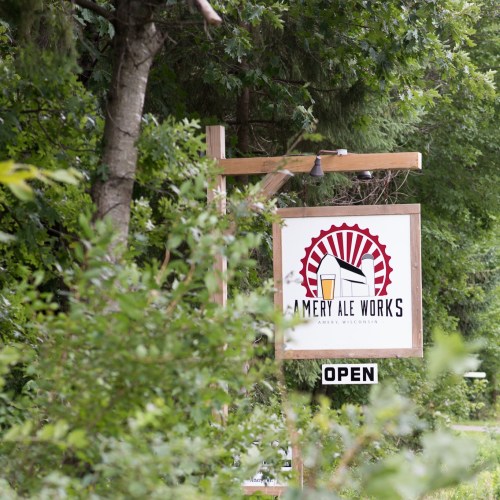 Sign for Amery Ale Works, featuring a logo with a beer glass and sunburst design, surrounded by greenery. "OPEN" sign visible.