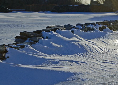A snow-covered stone wall with drifts of snow, set against a serene winter landscape.