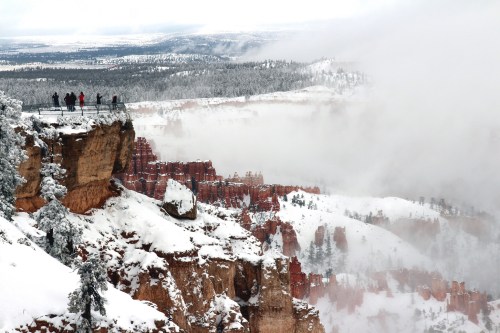 A snowy landscape with people standing on a cliff overlooking red rock formations and a misty valley.