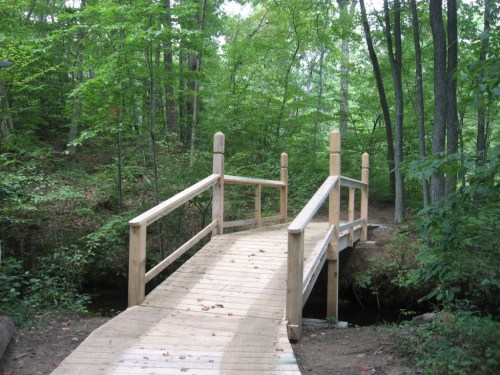 A wooden bridge spans a small creek, surrounded by lush green trees in a serene forest setting.