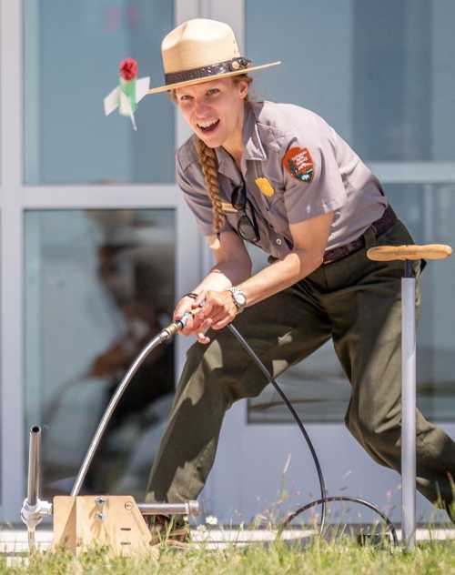 A park ranger in a hat demonstrates a science experiment with a device, smiling and engaged in the activity.