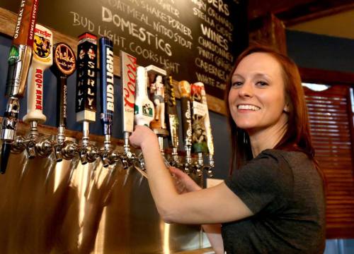 A smiling woman serves beer from a row of taps at a bar, with a chalkboard menu in the background.