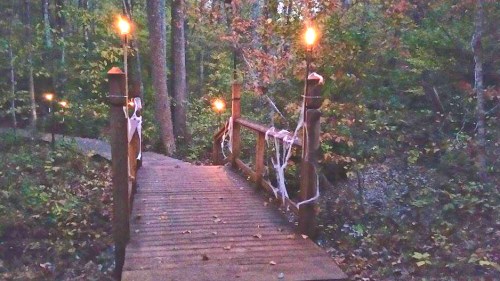 A wooden bridge in a forest, adorned with cobwebs and lit by torches, surrounded by autumn foliage.