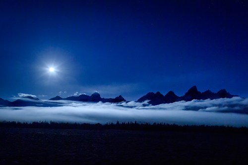 A serene night scene with a full moon illuminating mountains above a blanket of fog. Stars twinkle in the dark sky.