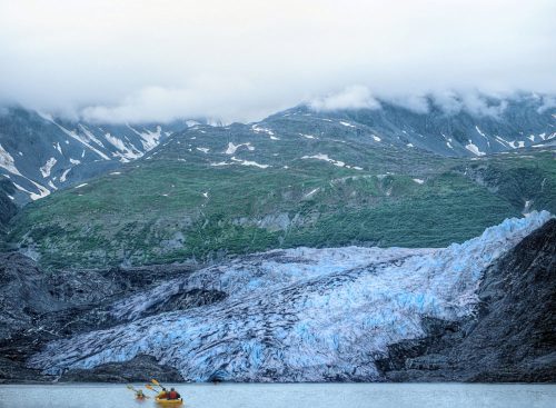A kayak approaches a massive glacier, surrounded by mountains and misty clouds in a serene landscape.