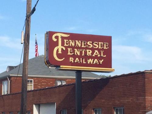Sign for Tennessee Central Railway, featuring bold lettering on a red background, with a building and flag in the background.