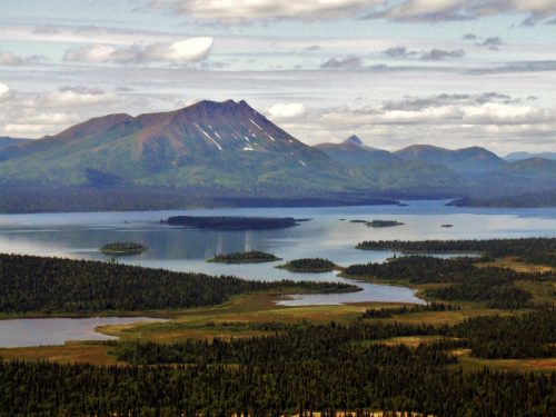 A scenic view of mountains and islands reflecting in a calm lake, surrounded by lush greenery and a cloudy sky.
