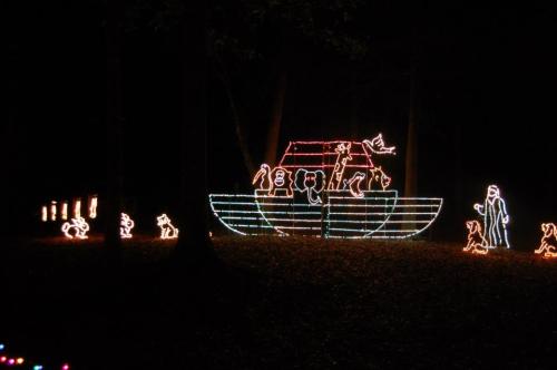 Colorful holiday lights depict a boat with figures and animals in a dark forest setting.