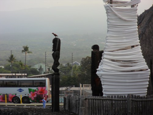 A totem pole stands beside a wrapped structure, with a bus and a person in blue in the background.