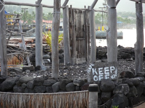 A weathered wooden structure with a "KEEP OFF" sign on a stone pillar, surrounded by rocky terrain and palm trees.