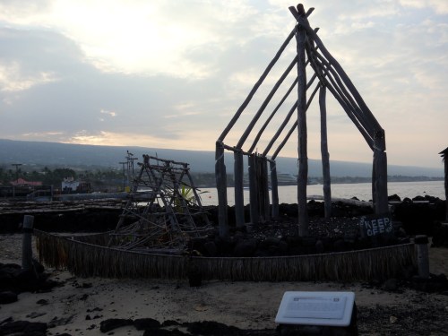 A partially constructed wooden structure on a beach, with a cloudy sky and water in the background.