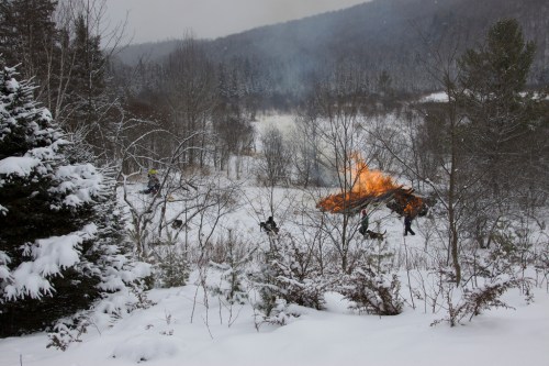 A snowy landscape with trees, where a fire burns in the distance, surrounded by a winter scene.