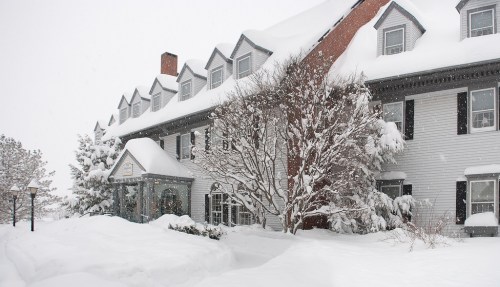 A snow-covered hotel with a large front porch, surrounded by trees and a winter landscape. Snowflakes are falling.