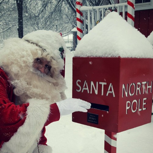 Santa Claus stands beside a snowy mailbox labeled "Santa North Pole," smiling and waving in a winter wonderland.