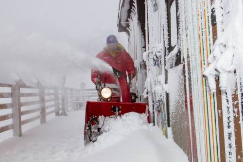 A person in a red jacket uses a snowblower to clear heavy snow near a building during a snowstorm.
